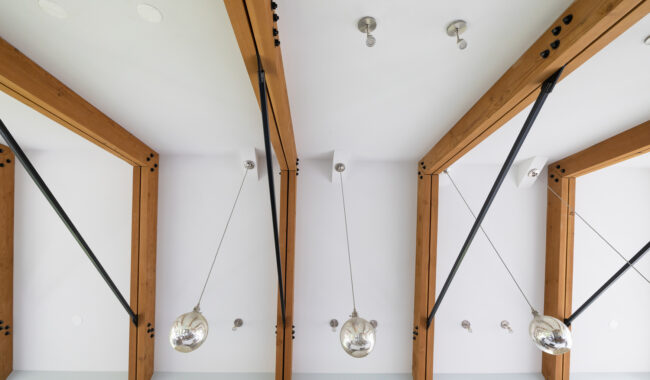 Ceiling view of a modern interior with wooden beams, black support rods, and three hanging glass pendant lights. The white ceiling is accented by exposed fixtures and hardware.