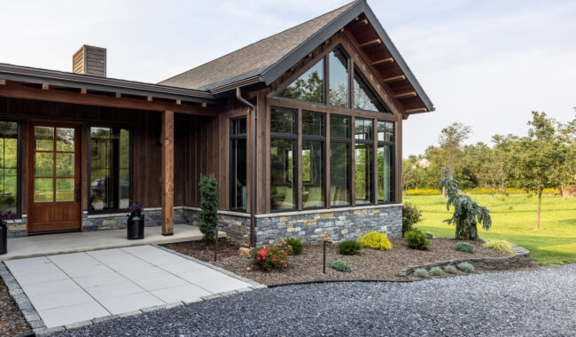 Modern house with large glass windows, wooden siding, and stone accents. The front features a paved walkway, landscaped garden beds, and a gravel driveway. Green grass and trees are visible in the background.
