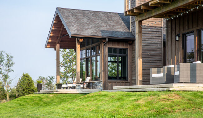 A modern wooden house with large windows and a covered porch, featuring outdoor seating on a concrete patio and surrounded by green grass and trees.