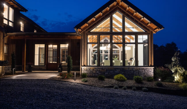 Modern house with large windows brightly lit from inside at dusk, showcasing exposed wooden beams and a dining area. The exterior includes stonework, plants, and a gravel driveway, with a deep blue evening sky in the background.