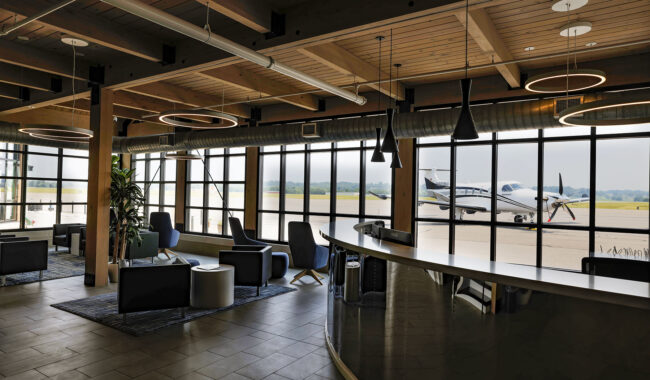 A modern airport lounge with contemporary seating and large windows, through which a small propeller airplane is visible parked outside on the runway. Warm lighting and wooden ceilings create a welcoming atmosphere.
