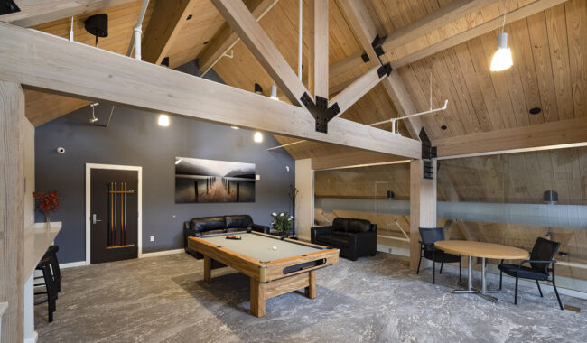 Spacious loft with exposed wooden beams, a pool table, black sofas, a round table with chairs, wall art, and modern lighting under a slanted wooden ceiling.