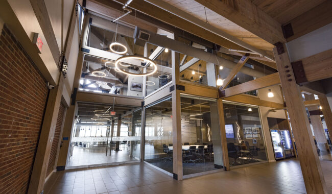 A modern office interior with exposed wooden beams, glass-walled meeting rooms, circular ceiling lights, tile flooring, and natural light coming in from large windows in the background.