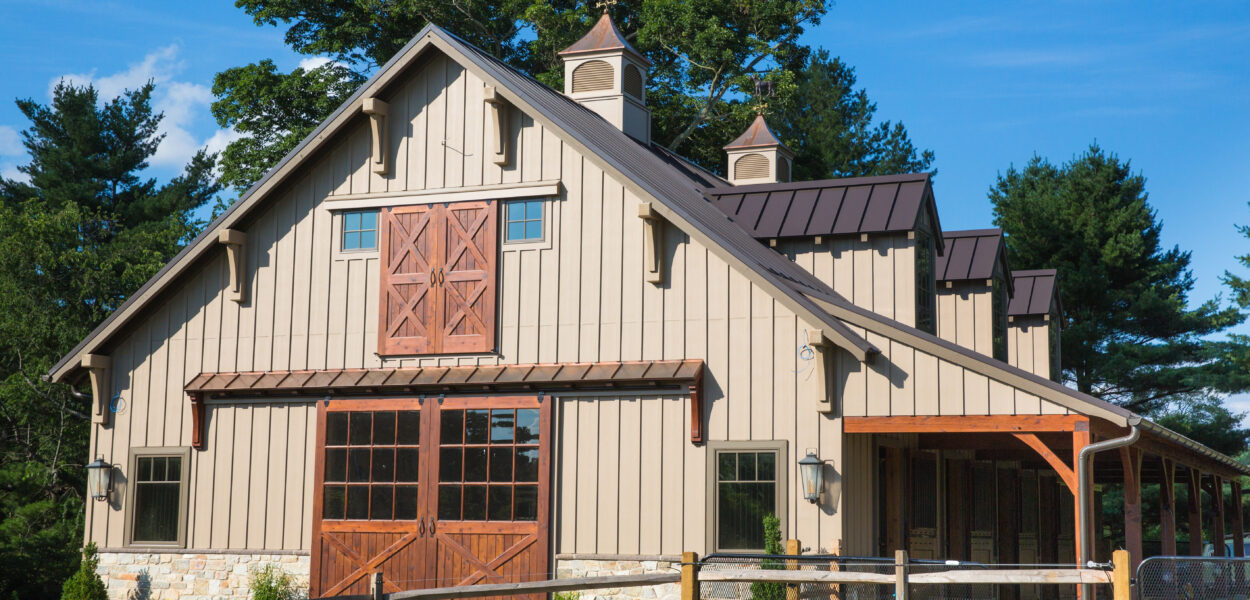 A large, beige barn with wooden doors and windows, cupolas on the roof, and a covered side area, surrounded by a wooden fence and trees under a clear blue sky.