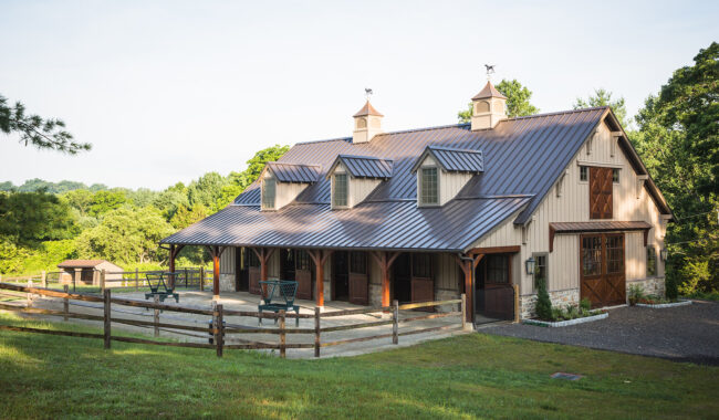 A large, modern barn with a metal roof, dormer windows, and cupolas sits in a green, rural area. The barn is surrounded by a wooden fence and trees, with chairs and tables on a concrete patio.