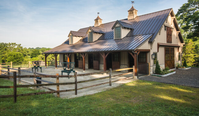 A large, tan barn with a dark metal roof stands on a sunny day, surrounded by green grass, a wooden fence, and shade trees. There are picnic tables outside, and the barn features multiple cupolas and windows.