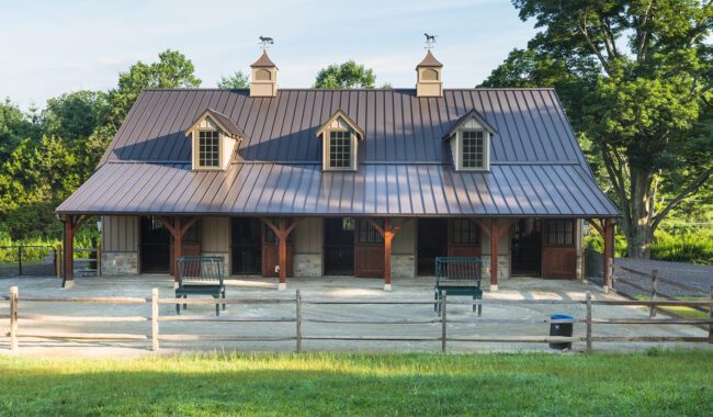 A large, brown horse barn with three dormer windows and cupolas topped with weather vanes, wooden doors, benches in front, and surrounded by green trees and grass under a clear sky.