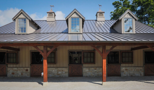 A barn with a metal roof, stone and wood walls, four dormer windows, and four wooden stable doors, set on a sunny day with blue sky and some trees in the background.