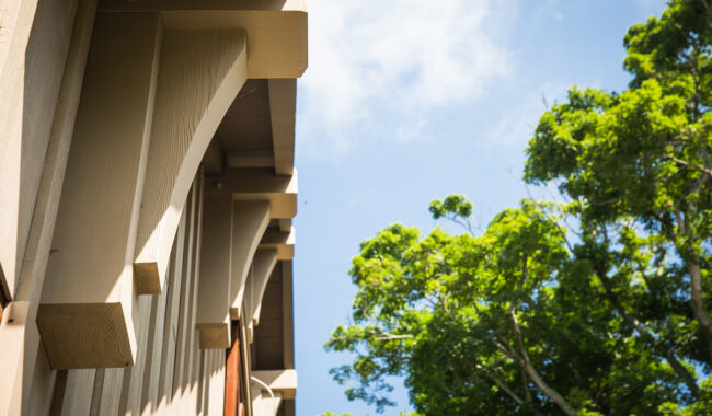 A close-up view of a wooden roof overhang with decorative brackets, set against a blue sky and tall green trees on a sunny day.