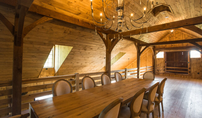 A rustic dining room with wooden walls, floors, and ceilings features a large wooden table surrounded by cushioned chairs. An elegant chandelier hangs above, and natural light streams through windows.