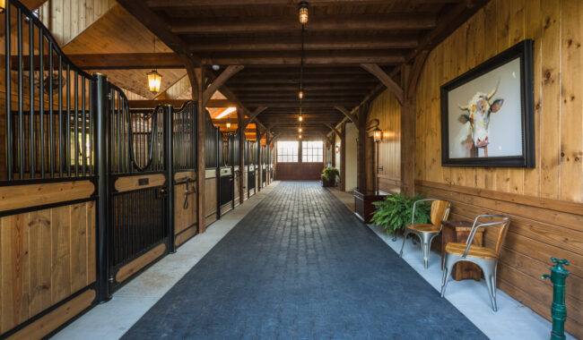 A neatly maintained horse stable with wooden walls, black metal stalls on the left, a paved central walkway, chairs and a painting on the right, and warm lighting throughout.