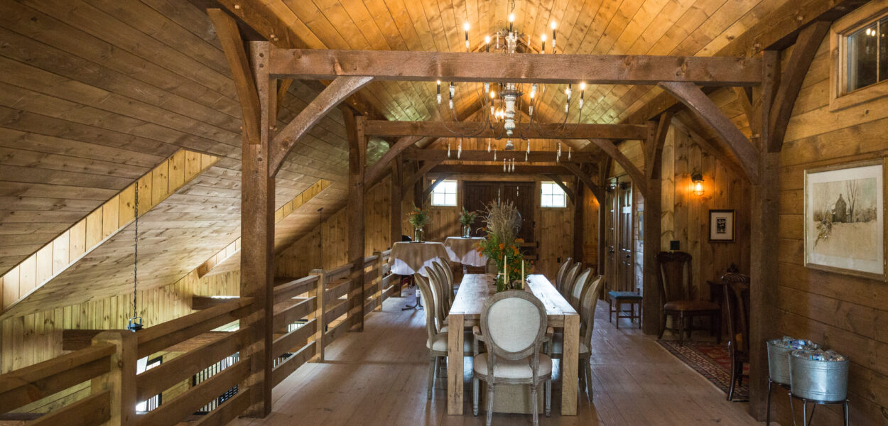 A rustic dining area with a long wooden table and upholstered chairs in a loft with wooden walls, ceiling, and floors. A chandelier hangs above, and natural light streams in through windows at the far end.