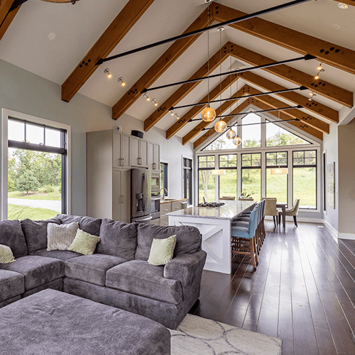 Open-concept living space with vaulted wood-beam ceiling, modern gray sectional sofa, white kitchen island with blue stools, dining area, large windows, and views of a green landscape outside.