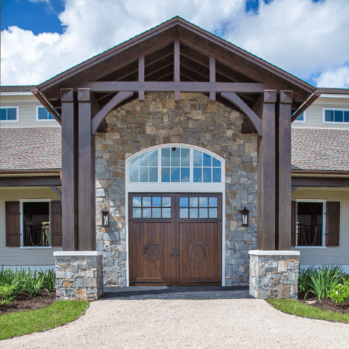 Large entryway of a modern house with stone and wood accents, double wooden front doors with window panes, tall wooden beams, and landscaped greenery along a gravel path.