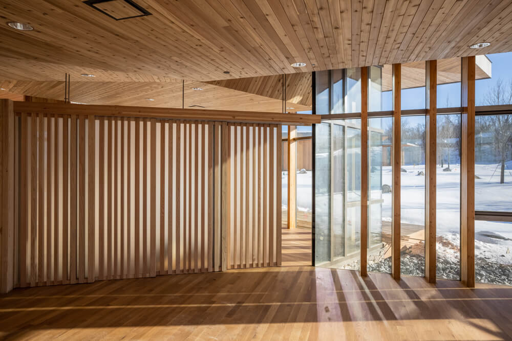 Modern interior with wooden floors, ceiling, and vertical slats in a stunning timber frame project gallery, large glass windows let in natural light with a snowy landscape visible outside.