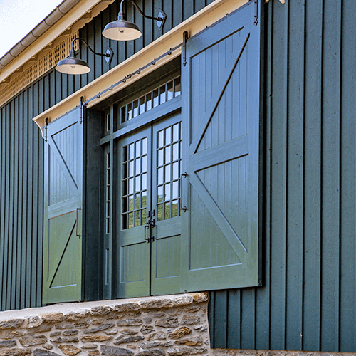 A green barn-style door with large shutters and glass window panes is set in a dark green wooden wall above a stone foundation, with three lights hanging overhead.