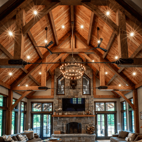A spacious living room with a high vaulted wooden ceiling, exposed beams, a large stone fireplace, a circular chandelier, modern ceiling fans, and large windows letting in natural light. Cozy seating is arranged below.