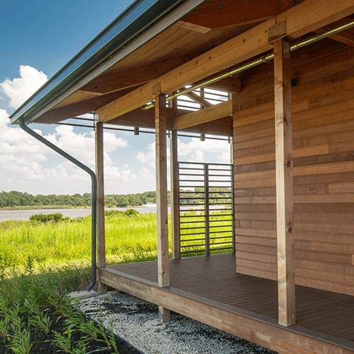 A modern wooden cabin with a covered porch overlooks a grassy field and a lake under a blue sky with scattered clouds.