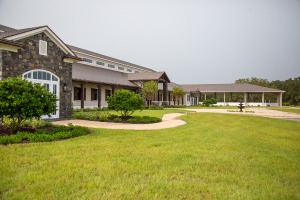 A large stone and wood building with a covered porch, surrounded by green grass, shrubs, and trees under a cloudy sky. A curved walkway leads to the entrance.