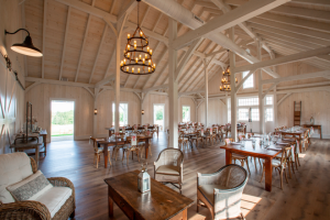 Spacious, rustic dining room with wooden floors, exposed beams, large windows, and chandeliers. Tables and chairs are arranged neatly, and natural light fills the room, creating a warm, inviting atmosphere.
