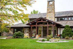A large stone and wood house with a covered front porch, tall stone chimney, manicured lawn, small bushes, and a gravel area in the front garden. Trees with green and yellow leaves surround the yard.