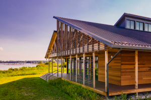 A modern wooden house with a slanted roof and large windows stands near a grassy area overlooking a body of water under a clear sky.