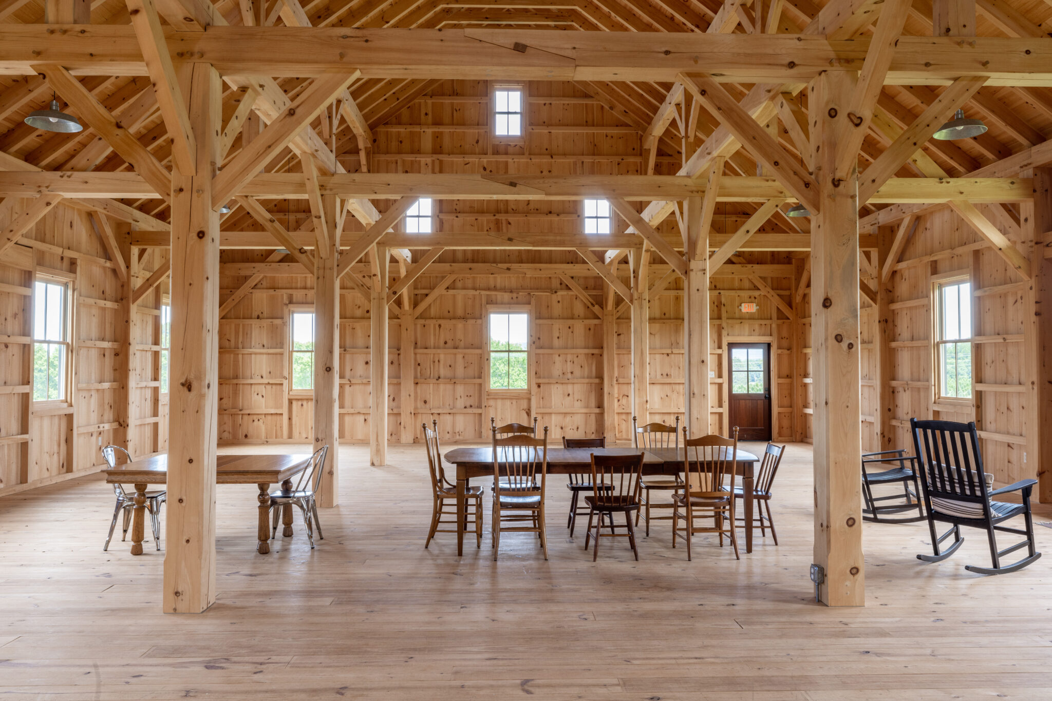 Spacious, sunlit wooden hall with exposed beams highlights this timber frame project gallery. Large windows illuminate dining tables and chairs, with two rocking chairs to the right and more tables on the left side of the room.