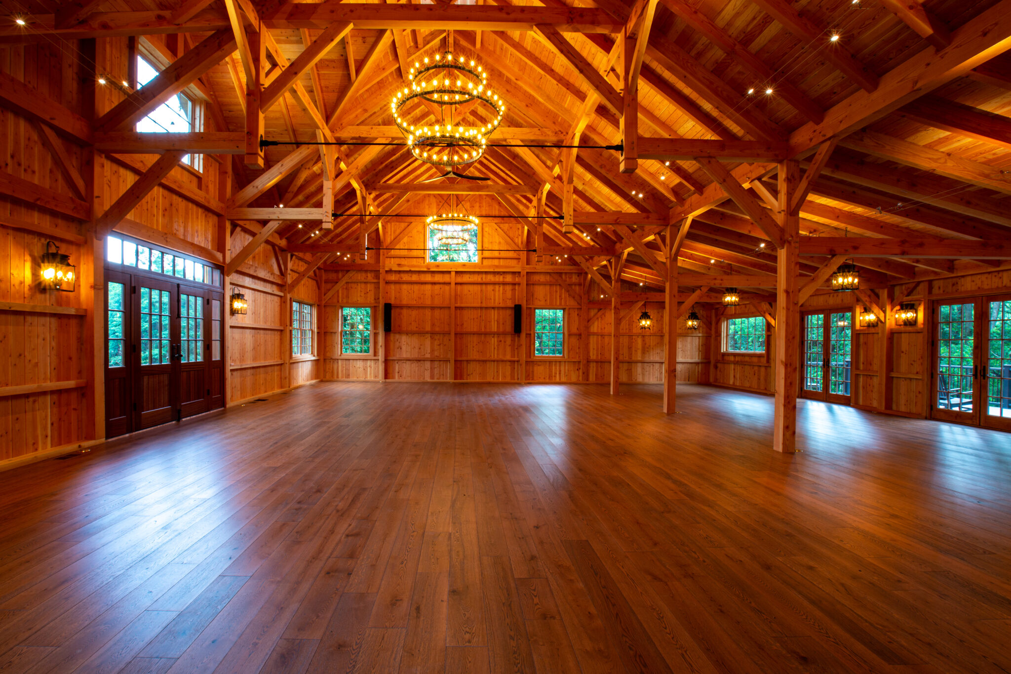 Spacious timber frame project gallery with a high vaulted ceiling, exposed beams, large windows, double doors, and elegant chandeliers creates a warm and inviting atmosphere with natural light streaming in.