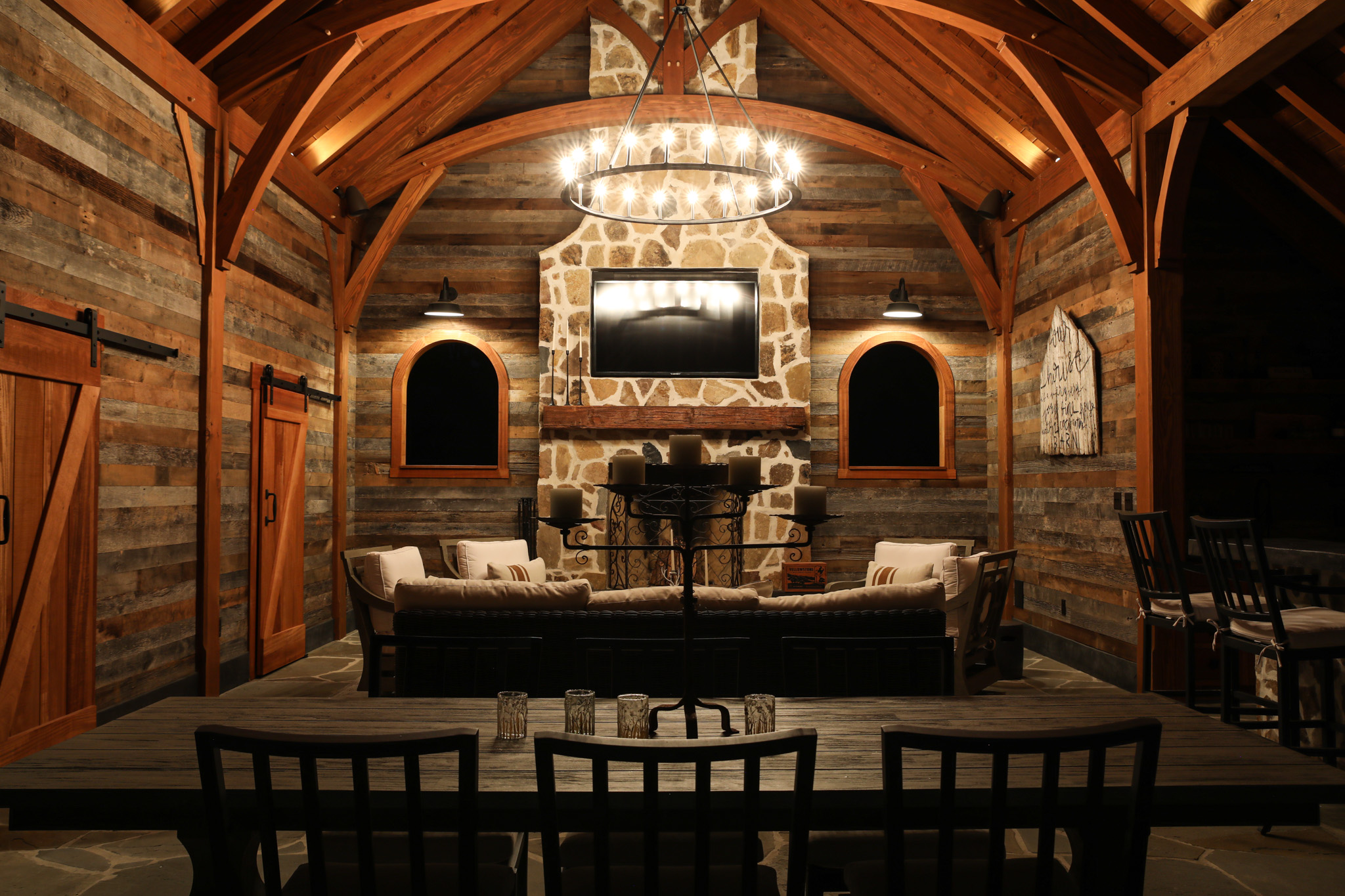 A rustic living room from our timber frame project gallery, featuring exposed wooden beams, wood-paneled walls, a stone fireplace, a circular chandelier, cozy sofas, and a wooden dining table with chairs in the foreground.