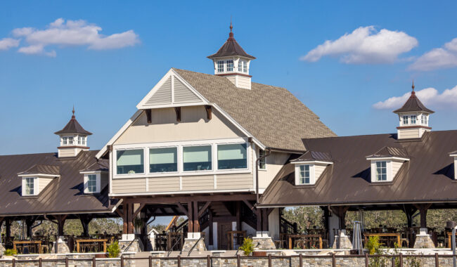 A large, modern barn with a gray metal roof and three cupolas under a blue sky with a few clouds, surrounded by a wooden fence and green grass.