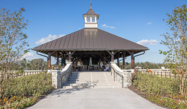 A large, open-sided pavilion with a metal roof and a cupola sits at the top of wide steps, surrounded by landscaped flower beds and trees under a clear blue sky.