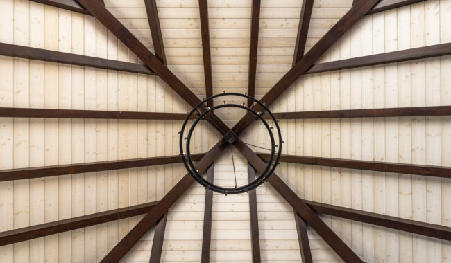 A view looking up at a wooden domed ceiling with dark beams radiating outward and a circular metal chandelier hanging in the center. The ceiling panels are light-colored wood.
