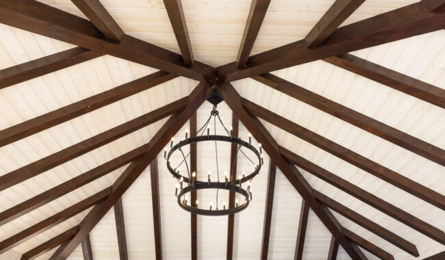A circular black chandelier with candle-style lights hangs from the center of a vaulted ceiling with dark wooden beams and white wooden panels.