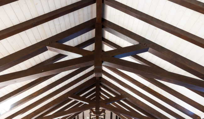 View of a ceiling with exposed, dark wooden beams arranged in a geometric pattern against a white wood-paneled roof, creating a symmetrical and architectural design.