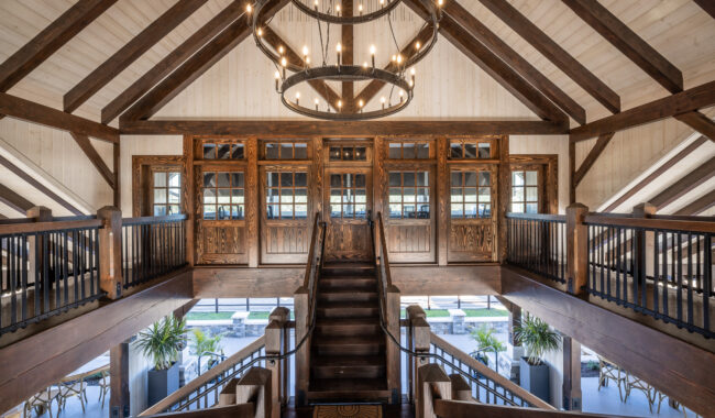 Grand wooden staircase with dark railings leads to a set of ornate double doors and windows under exposed beams, with a large circular chandelier hanging from the ceiling and plants visible below.