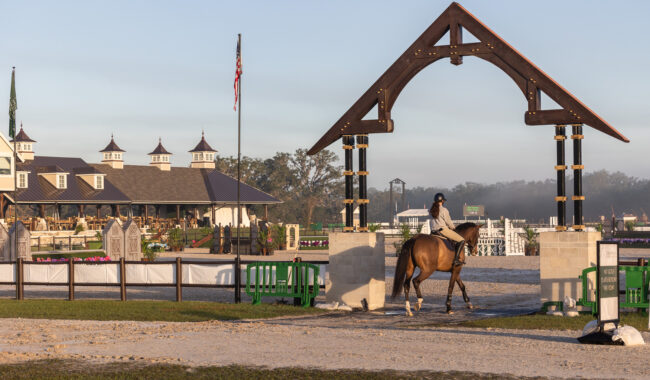 A person riding a brown horse passes under a large wooden arch at an equestrian facility, with a building, flags, fences, and green benches in the background on a clear day.