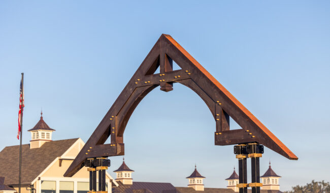 A close-up of a wooden triangular archway structure with metal supports, set outdoors against a clear blue sky. In the background, there is a building with multiple cupolas and an American flag.