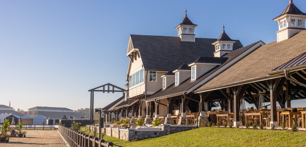 A large, elegant barn-style building with cupolas, outdoor seating, and a fenced riding area under a clear blue sky, surrounded by grass and landscaping.