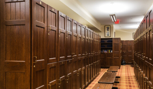 A well-lit locker room with rows of wooden lockers, a long wooden bench in the center, and a patterned carpet floor. Shelves with toiletries and a black bag are visible in the background.