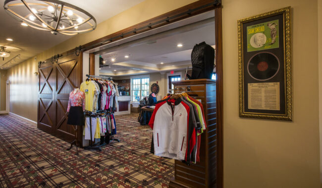Golf pro shop interior with racks of colorful shirts and jackets near a large wooden sliding door. Framed record and article hang on the beige wall; patterned carpet covers the floor under warm ceiling lights.