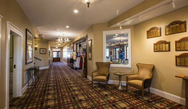 A hallway with plaid carpet, beige walls, chairs with a small table, wall plaques, a window into another room, and a display of clothing near glass doors at the far end. Warm lighting creates a welcoming atmosphere.