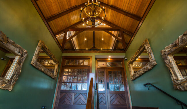 A rustic entryway with green walls, large wooden doors, ornate wood-framed mirrors on each side, a wooden ceiling with exposed beams, and a chandelier hanging above.