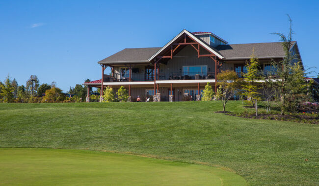 A large, two-story clubhouse with a sloped roof overlooks a well-manicured golf course, with green grass and trees under a clear blue sky.