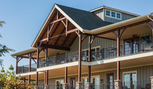 Two-story building with a steep, gabled roof, wooden beams, stone columns, and a large balcony featuring outdoor seating. The exterior is gray with white trim, and the sky is clear and blue.