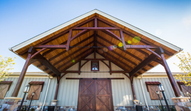 A wooden building with a high peaked roof and large wooden double doors; stone accents at the base and small lanterns line the entrance on a clear sunny day.