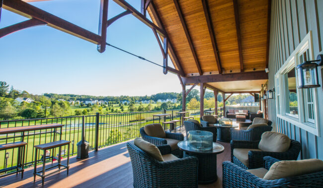 A covered outdoor patio with wicker chairs and glass tables overlooks a golf course and green landscape under a clear blue sky. The patio has a vaulted wooden ceiling and black metal railing.