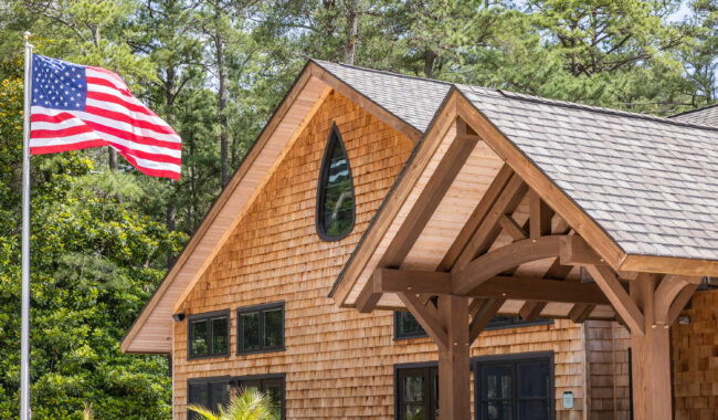 A wooden house with shingle siding and dark-framed windows sits among trees, with an American flag waving on a flagpole in the foreground. The sky is clear and sunny.