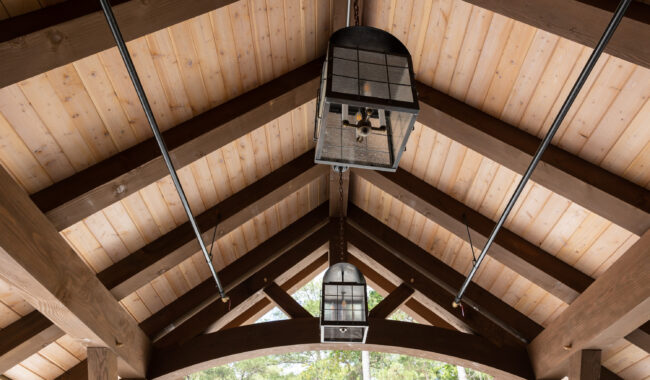 Wooden ceiling with exposed beams, metal rods, and two hanging lantern-style light fixtures, seen from below with trees visible through the openings at the sides.