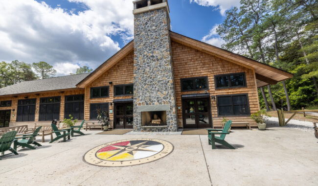 A rustic lodge with a tall stone chimney stands behind a courtyard featuring a large compass rose design. Green Adirondack chairs surround the area, and the building is framed by trees under a partly cloudy sky.