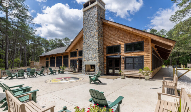 A large lodge with a stone chimney and wood siding sits among tall trees, with green Adirondack chairs arranged on a spacious patio and a compass design in the center under a partly cloudy sky.